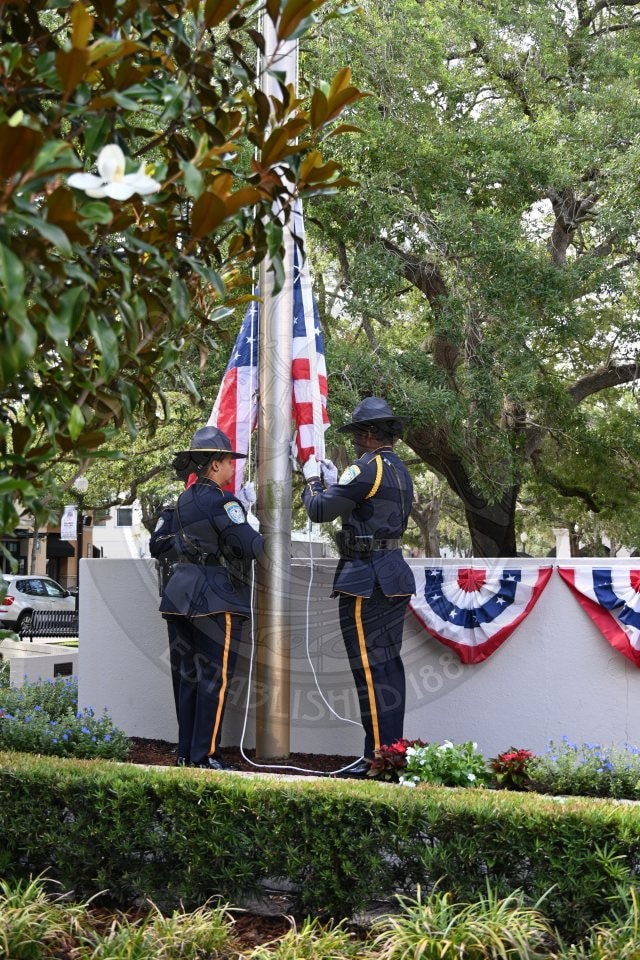 Two police officers raise an American flag on a flagpole beside a wall decorated with patriotic bunting.