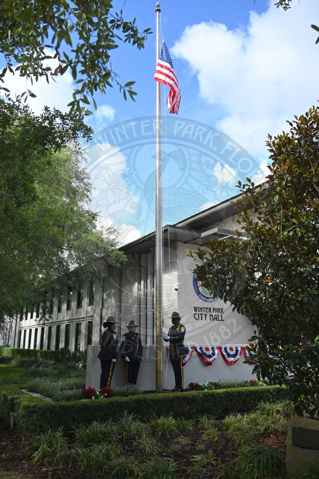 Four uniformed officers stand at attention by a flagpole outside Winter Park City Hall, with the U.S. flag raised.