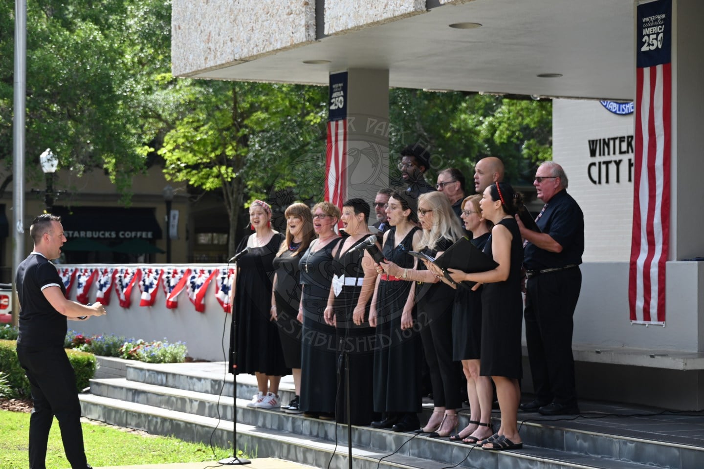 A choir sings outdoors on a decorated stage while a conductor leads them near a city hall building.