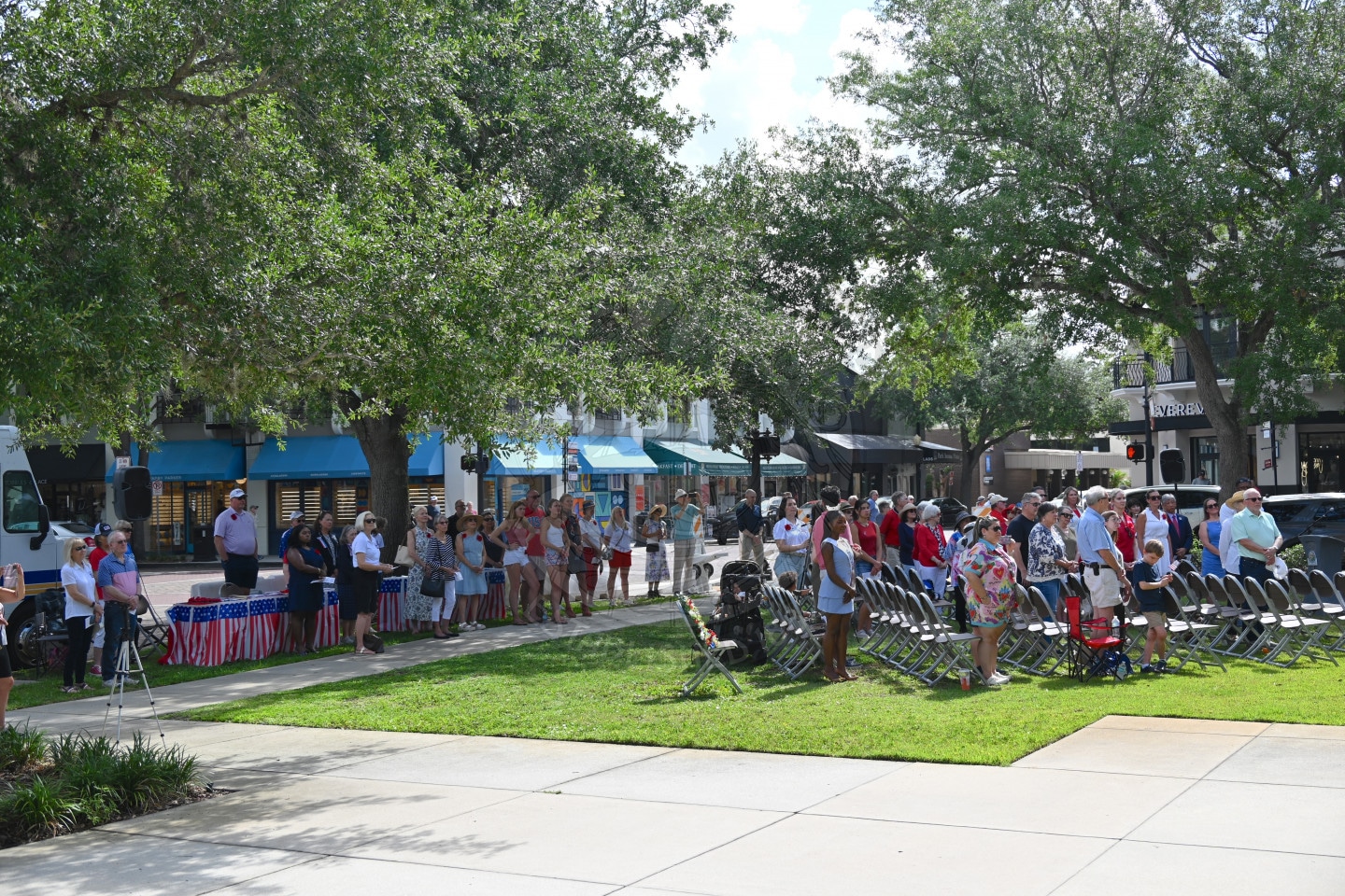 A group of people gather outdoors, some standing, some seated, surrounded by trees and buildings on a sunny day.