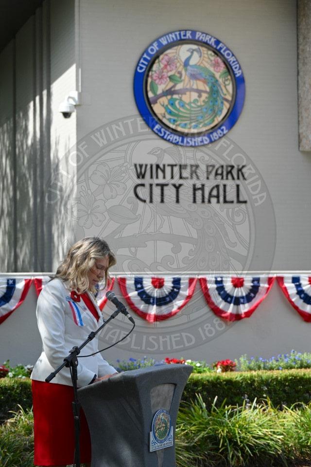 A woman speaks at a podium outside Winter Park City Hall, decorated with patriotic red, white, and blue bunting.