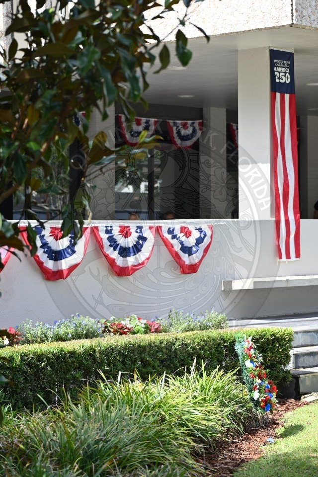 Red, white, and blue bunting and flag decorations on a building for a patriotic celebration; greenery in the foreground.