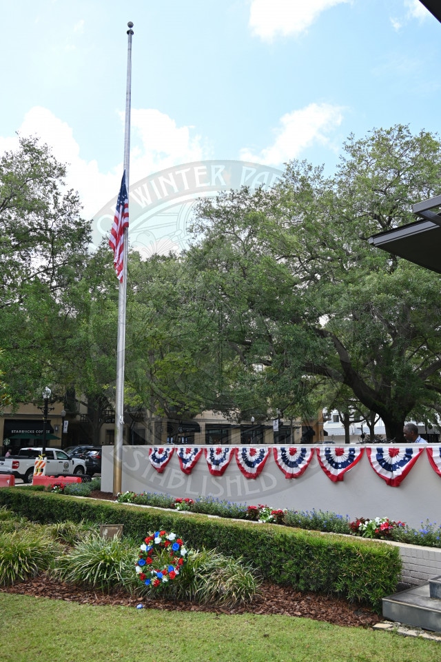 U.S. flag at half-staff, red, white, and blue bunting, and wreath in front of a memorial on a sunny day.