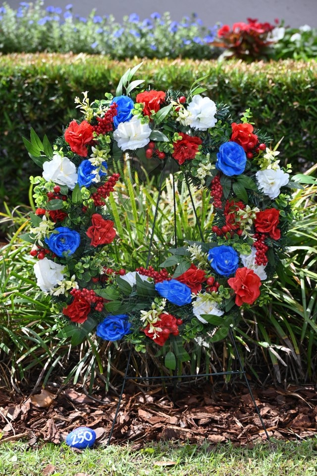 A wreath of red, white, and blue flowers on a stand is displayed outdoors, surrounded by greenery.