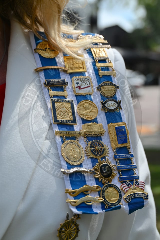 Close-up of a womans shoulder displaying various gold and blue medals and pins on a white and blue ribbon sash.