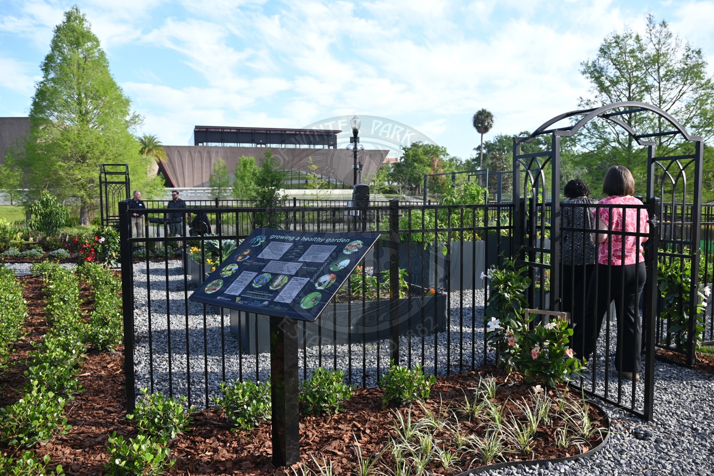 A garden with an informational sign, black fence, and two women standing at a gate, with a building in the background.