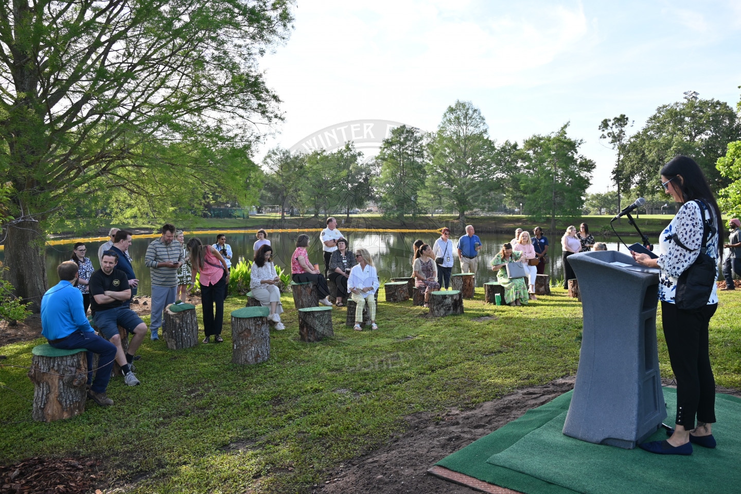 A woman speaks at a podium outdoors to a group seated on tree stumps by a lake surrounded by trees.