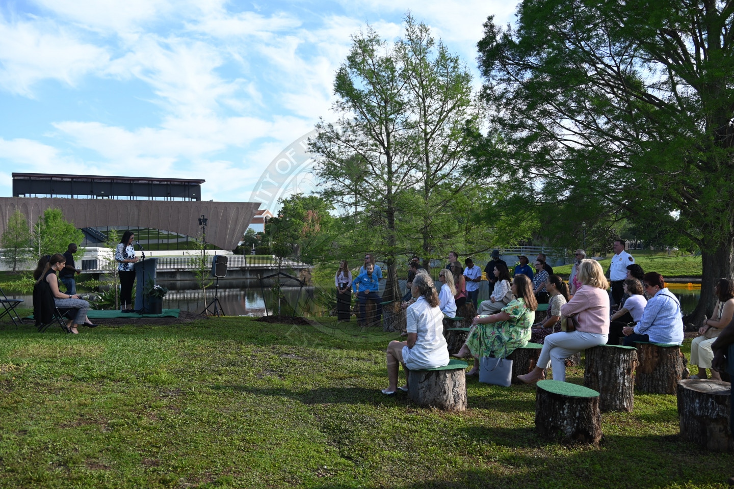 A group of people sit on tree stumps outdoors, listening to a speaker near a pond with trees and a building in the background.