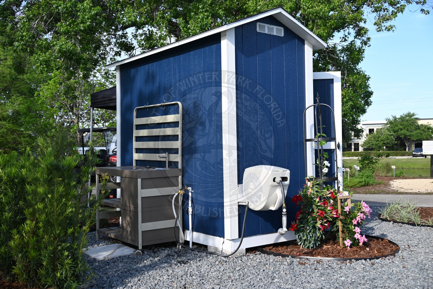 A small blue shed with white trim sits on gravel, beside a water spigot and blooming pink flowers.