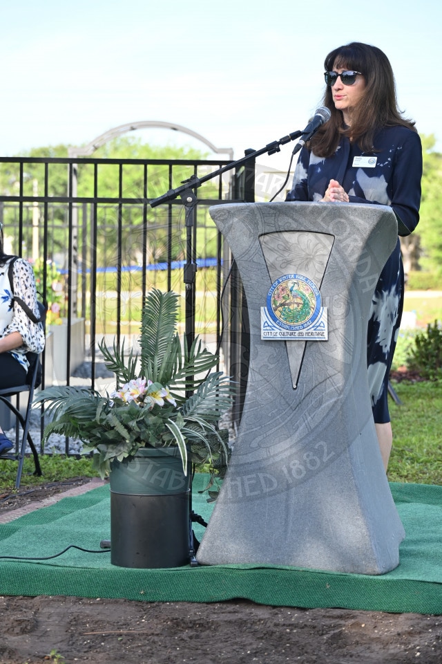 A woman in sunglasses speaks at an outdoor podium with a plant and city seal, during a daytime event.
