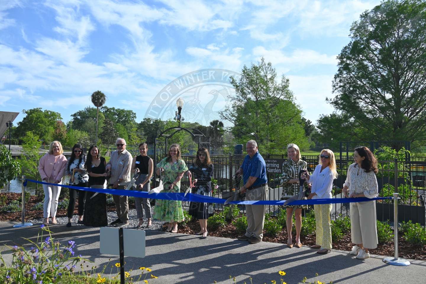 A group of people stand behind a blue ribbon outdoors, preparing for a ribbon-cutting ceremony at a garden.
