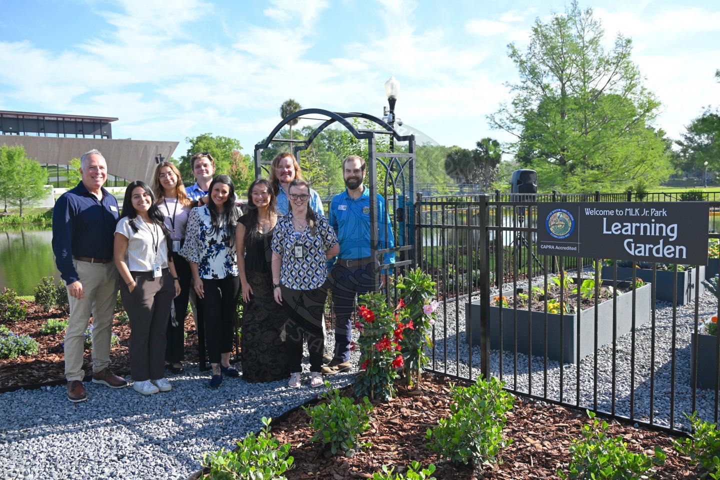 A group of people stand smiling by a sign that reads Learning Garden in a sunny outdoor garden setting.