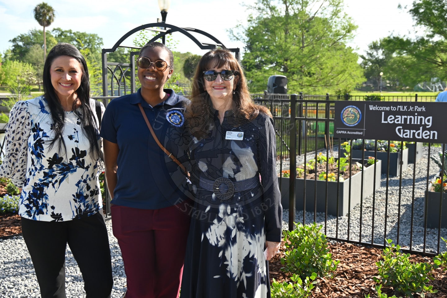 Three women stand smiling in front of a sign that reads Learning Garden at a sunny outdoor garden area.