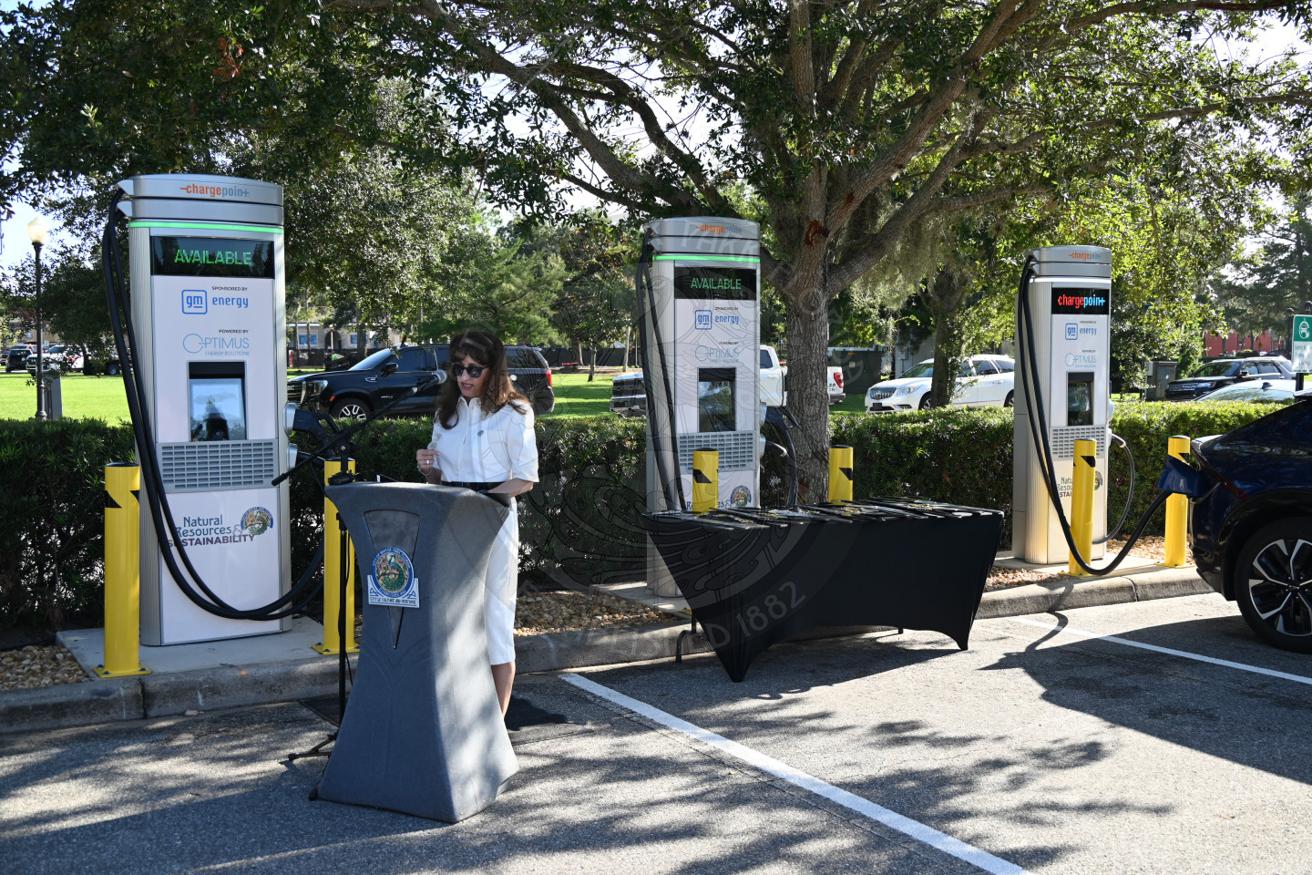 a lady standing behind a podium speaking into a microphone with ev charging stations behind her.