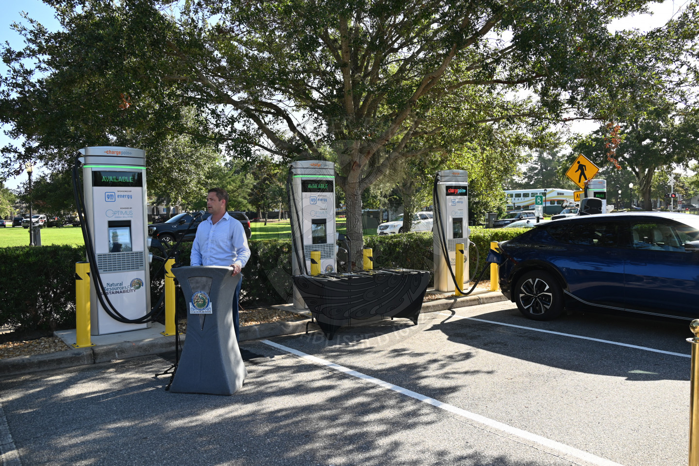 a man standing behind a podium speaking into a microphone with ev charging stations behind her.