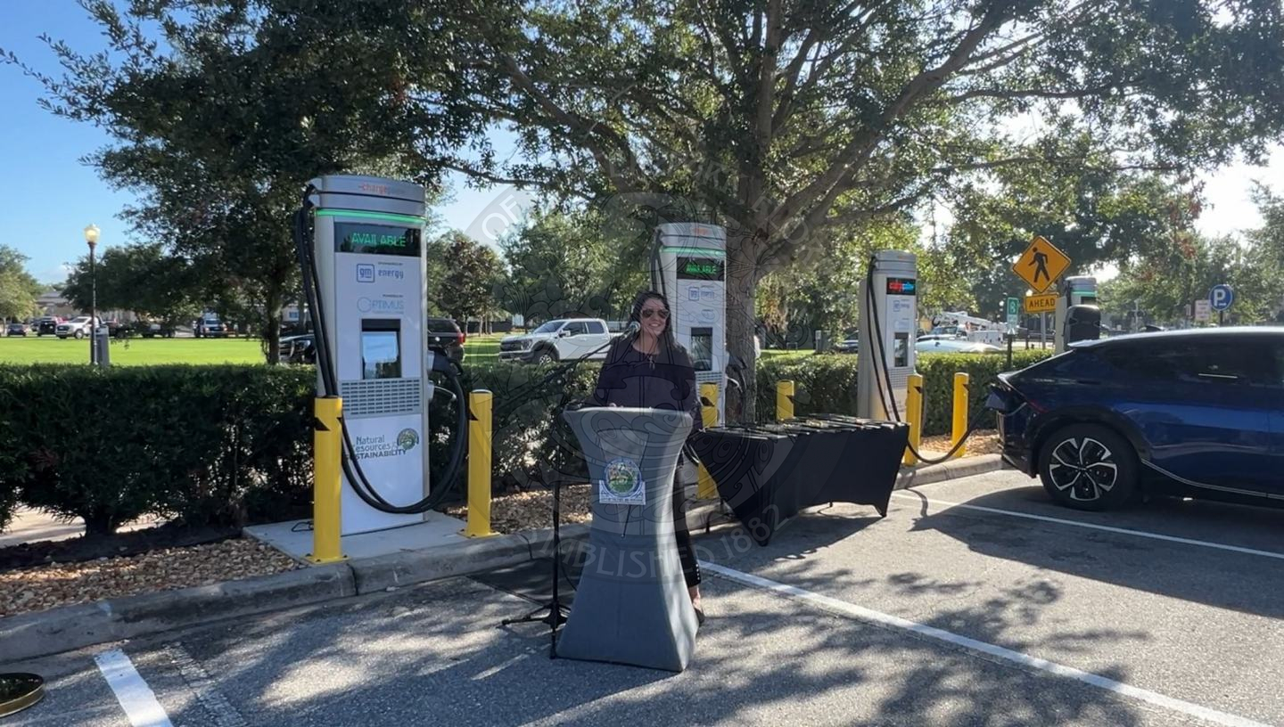 a lady standing behind a podium speaking into a microphone with ev charging stations behind her.