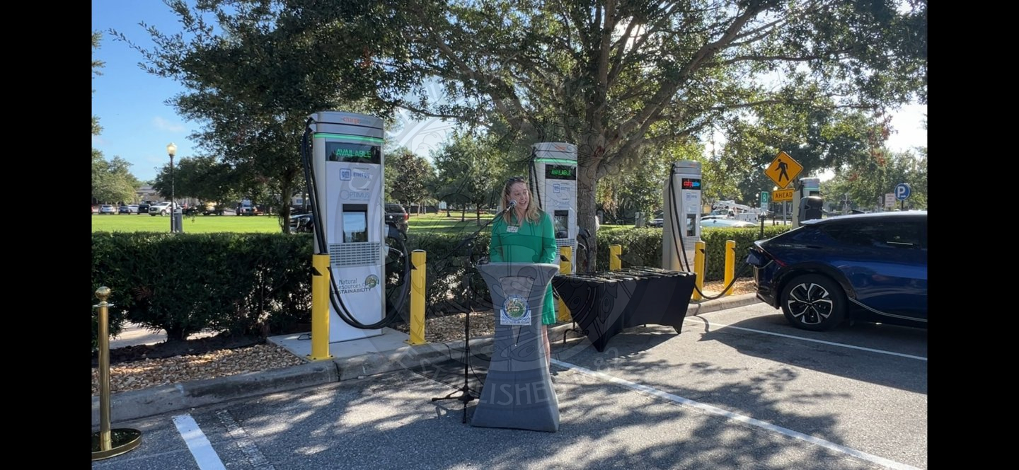 a lady standing behind a podium speaking into a microphone with ev charging stations behind her.