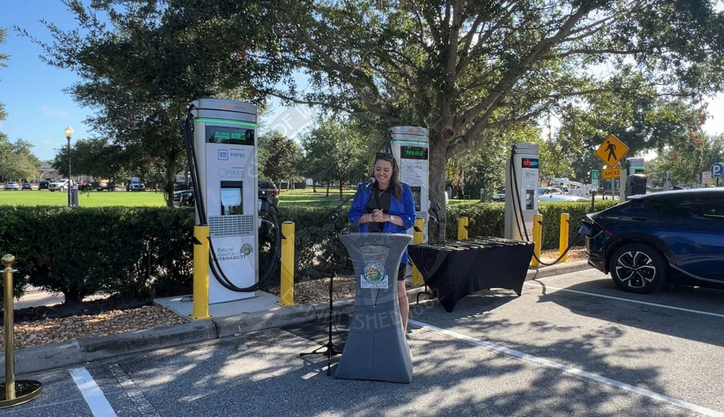 a lady standing behind a podium speaking into a microphone with ev charging stations behind her.