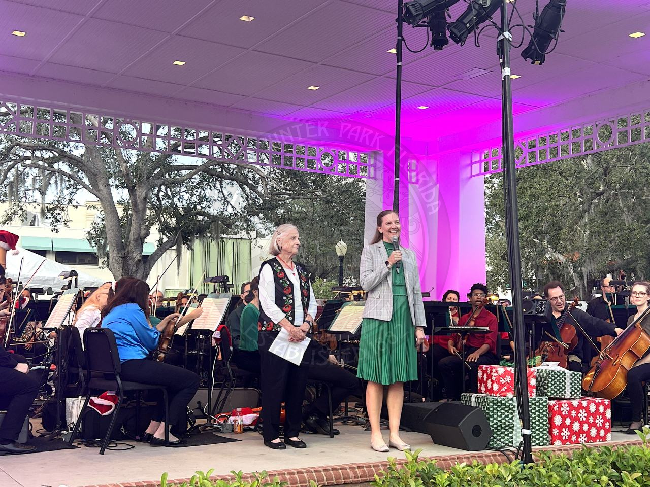 two women standing on the stage facing the crowd of people sitting in lawn chairs. One women is holding a microphone and talking and the other one is standing next to her holding a peice of paper. 