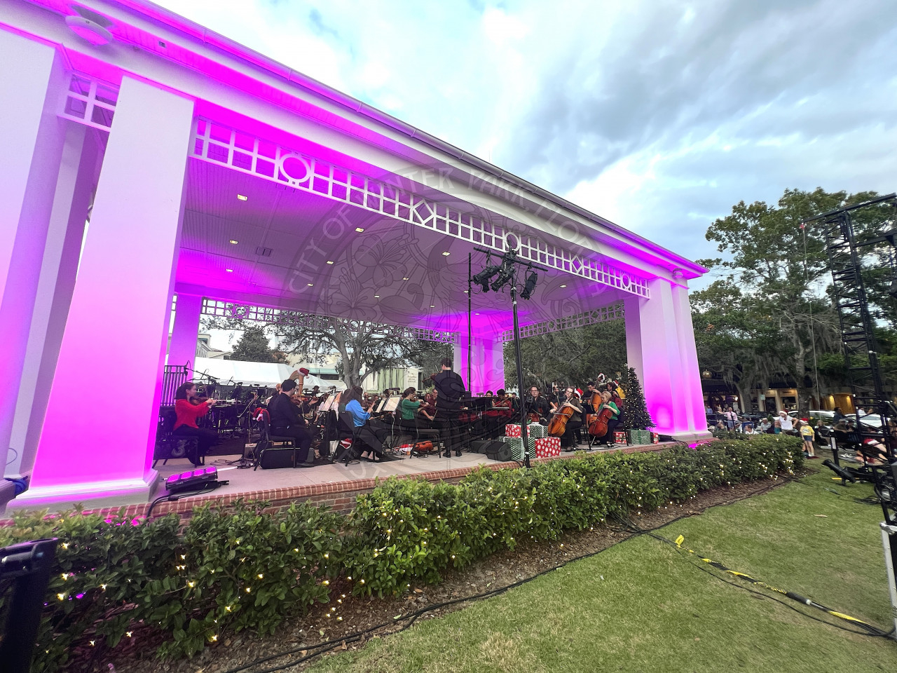 Man standing in front of the stage with his back turned to the crowd conducting a philharmonic orchestra with pink lights on the stage.