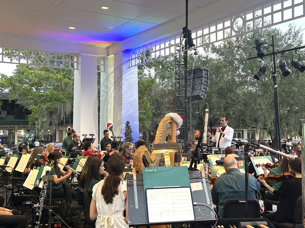 a view from behind the stage with the philharmonic orchestra playing and one man standing conducting the page. You can see the crowd watching them.