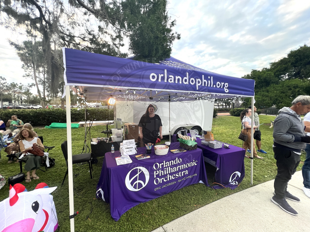 a 10x10 tent with 3 tables underneath and purple linens that say Orlando Philharmonic Orchestra. There is a lady behind the front table standing and smiling at people.