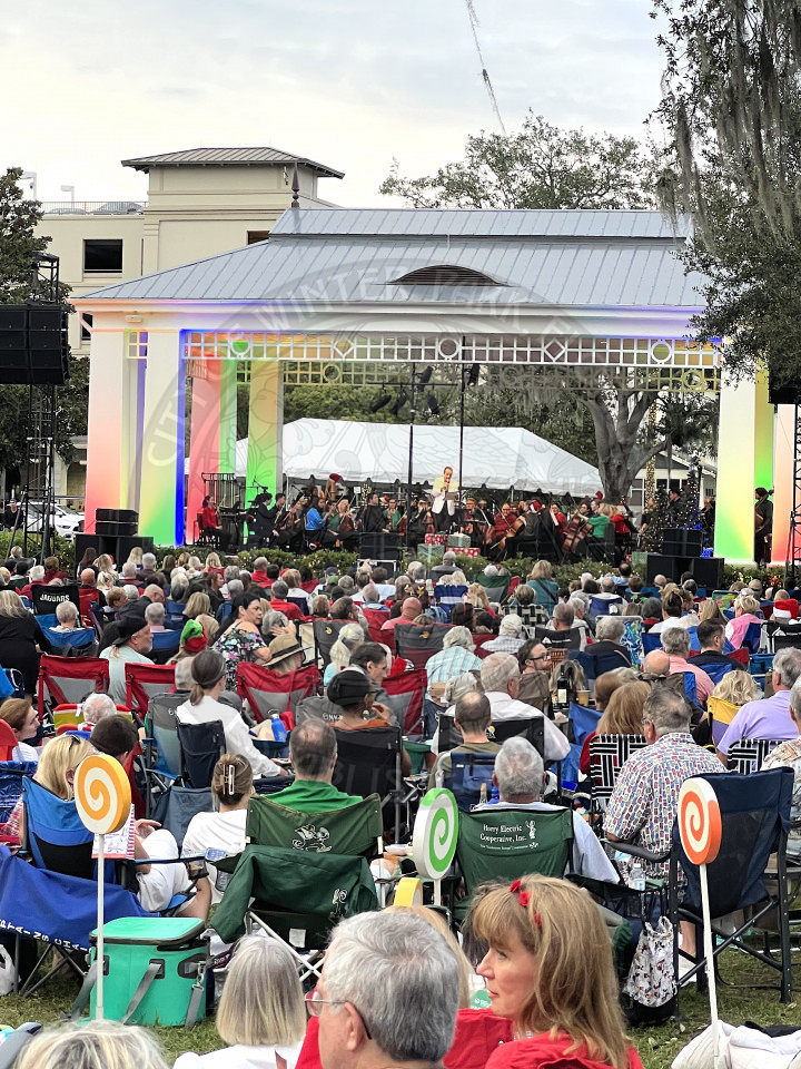 Crowd of people sitting in lawn chairs facing a stage with the Orlando Philharmonic Orchestra on the stage getting ready to perform. There is a man in the middle of the stage talking to the crowd. 