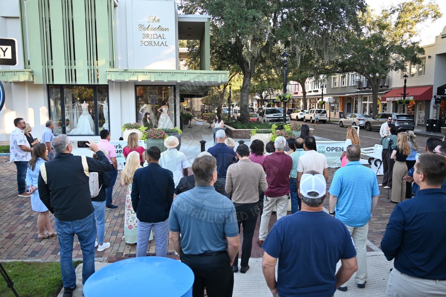 A crowd of people stands on a brick street facing the front entrance of a bridal boutique.