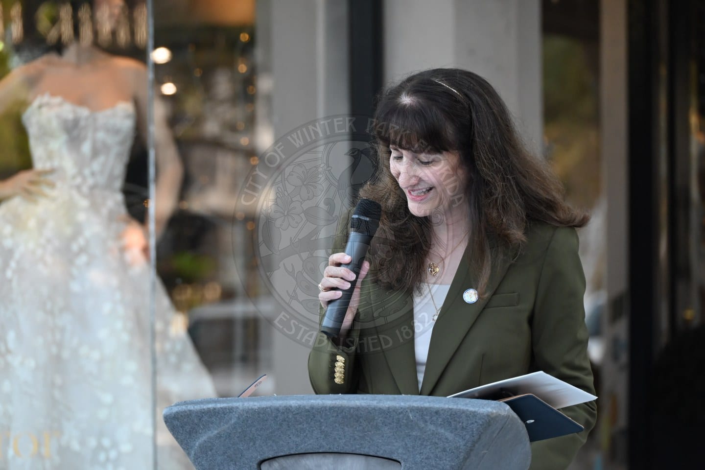 A woman in an olive green blazer speaks into a microphone at a podium.