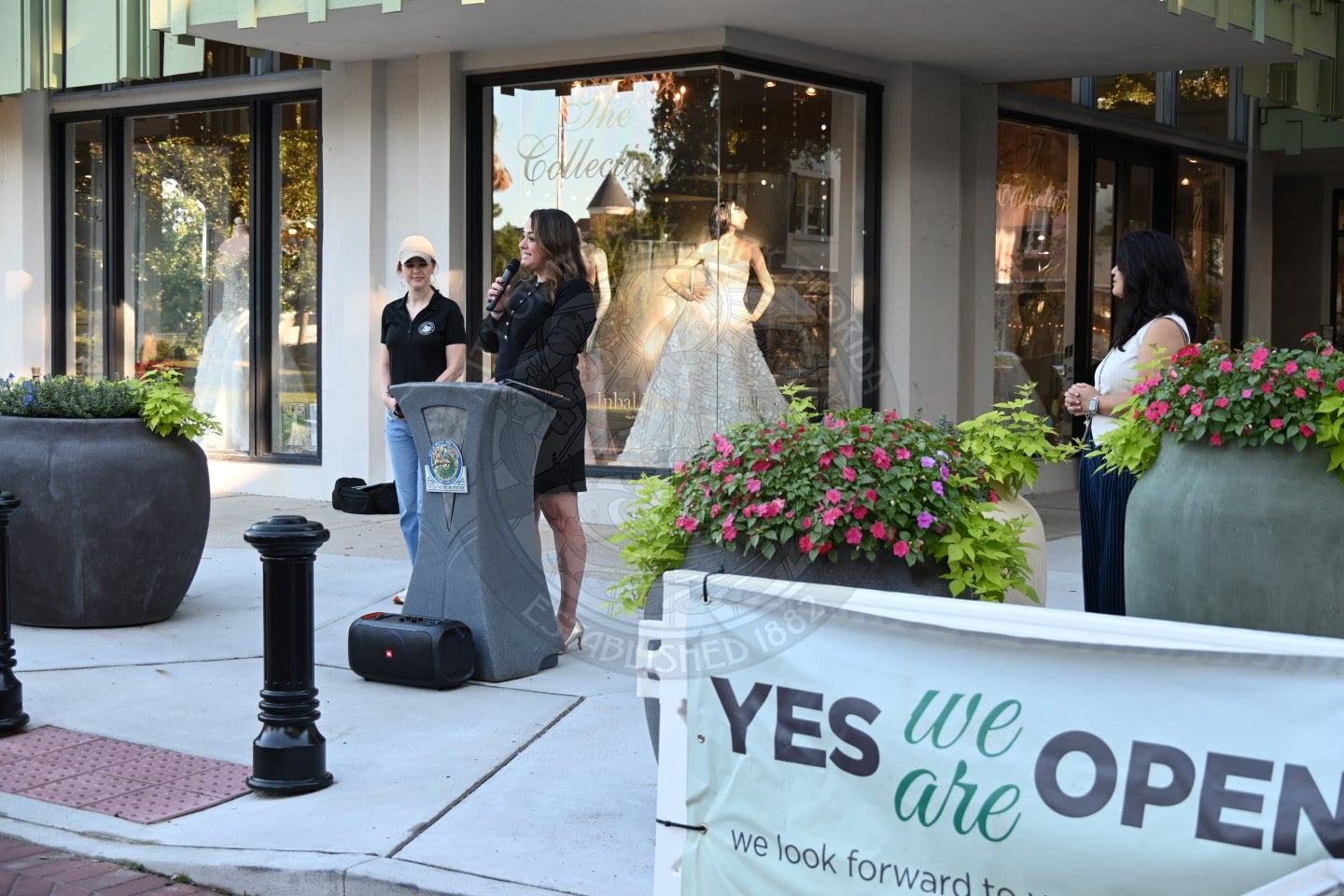 A woman speaks into a microphone at a podium outside a bridal shop with a YES we are OPEN sign in the foreground.