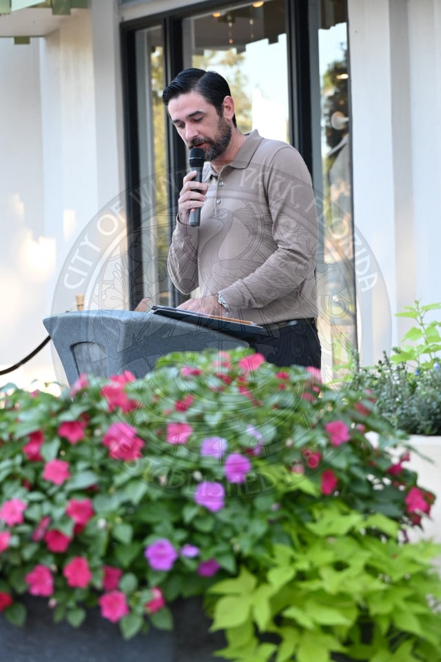 A man in a tan long-sleeved polo shirt speaks into a microphone from behind a podium.