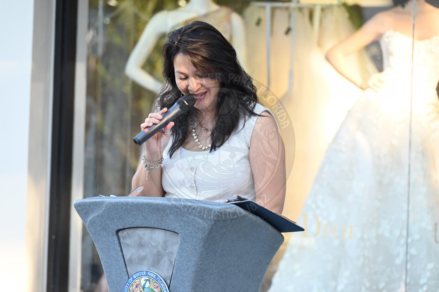 A woman speaking into a microphone from a podium in front of a wedding dress display.