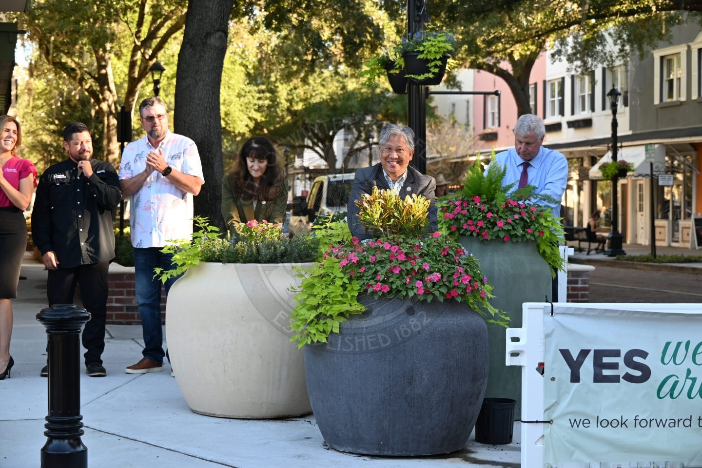 A group of people standing outdoors near large planters filled with pink and green flowers.