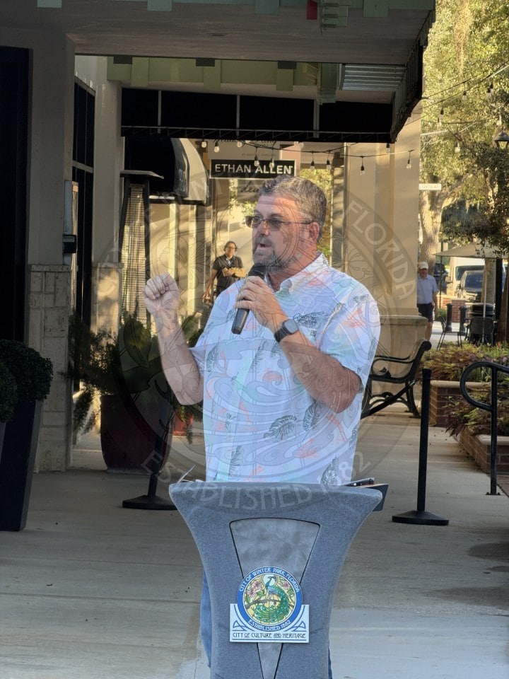 A man in a patterned button-down shirt speaks into a microphone at a podium with the Winter Park, Florida city seal.