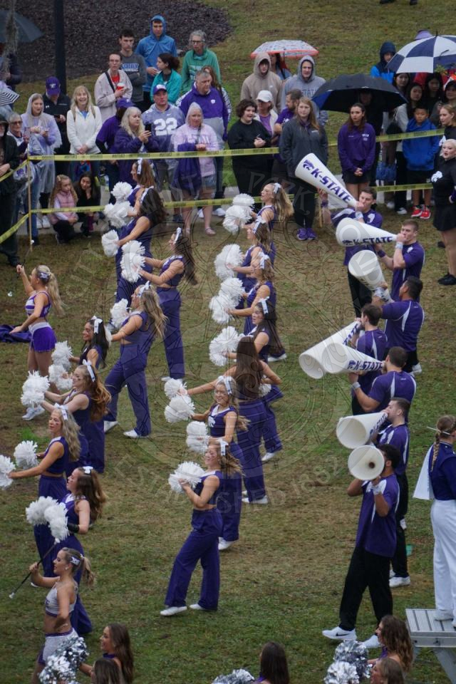 Kansas State Cheerleaders chanting in front of a crowd. The female cheerleaders are clapping their hands with pom poms and the male cheerleaders that are behind them  are shouting throguh plastic cheer megaphones.