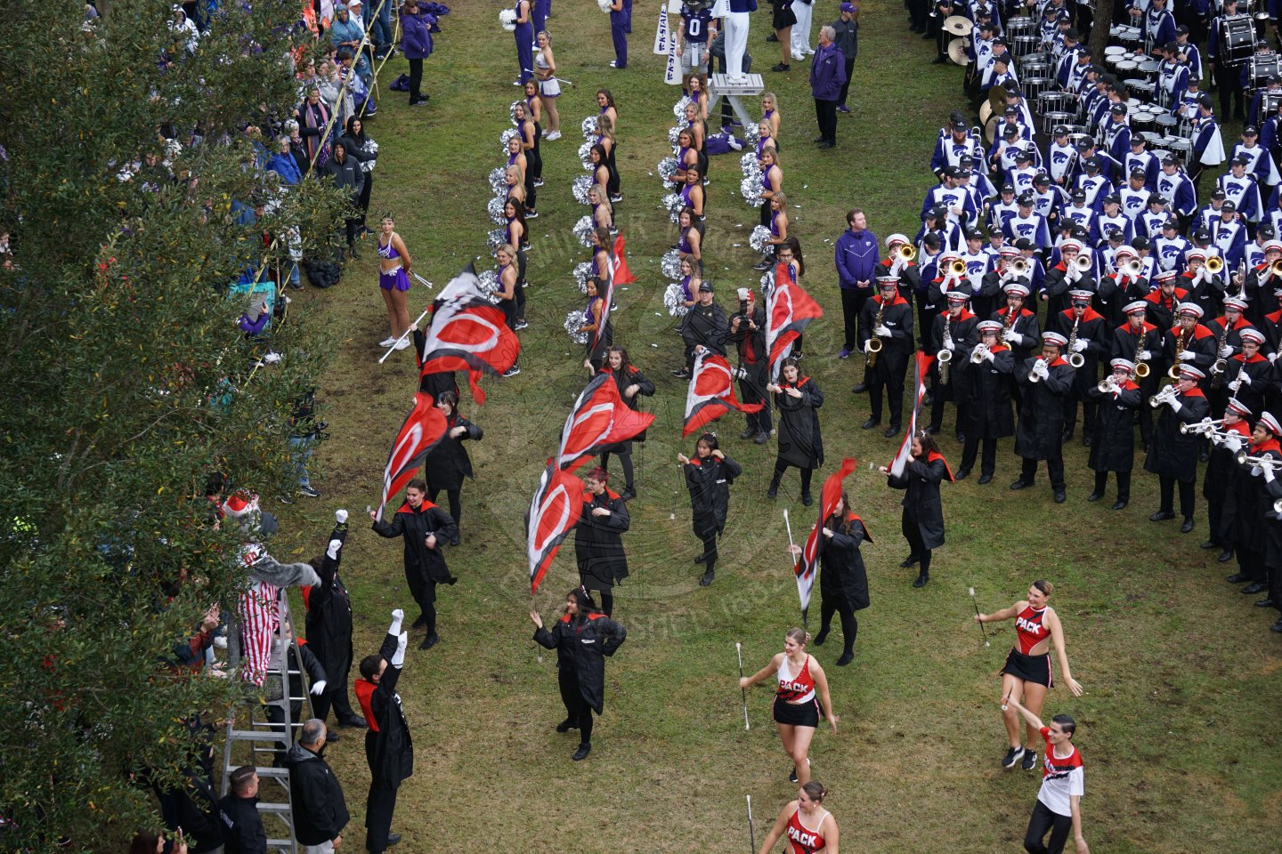 Two college bands standing in an arch with cheereleaders, dancers, and colorguard members standing in front of th e arch. One band is playing their insturment and the colorguard members are using the flags to cooridnate with the music.