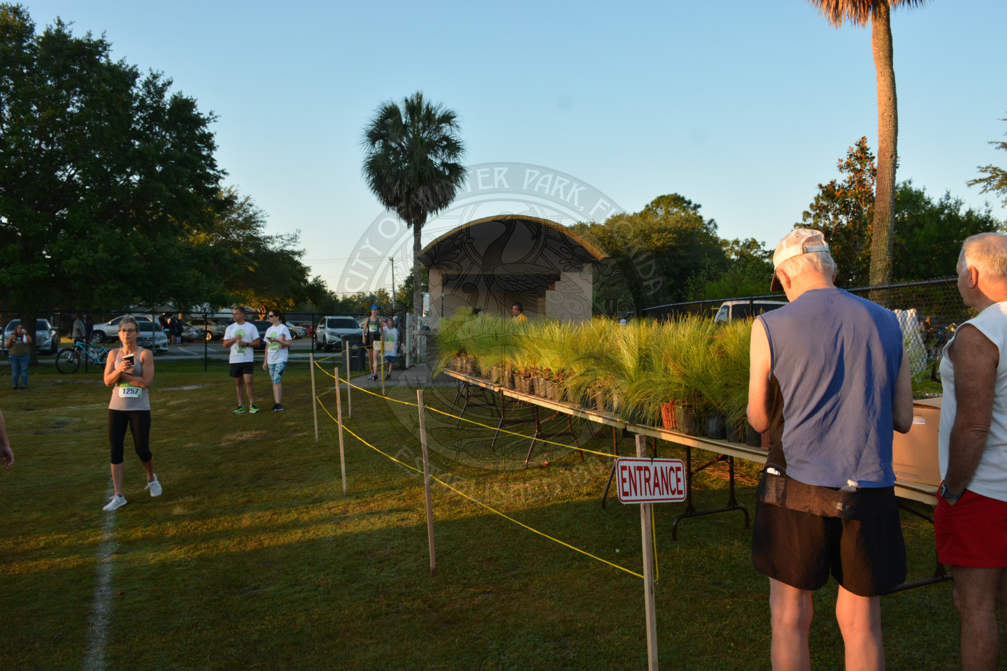 Two males standing at entrance sign, one lady waiting in the grass and an empty cue line with potted plants on a table