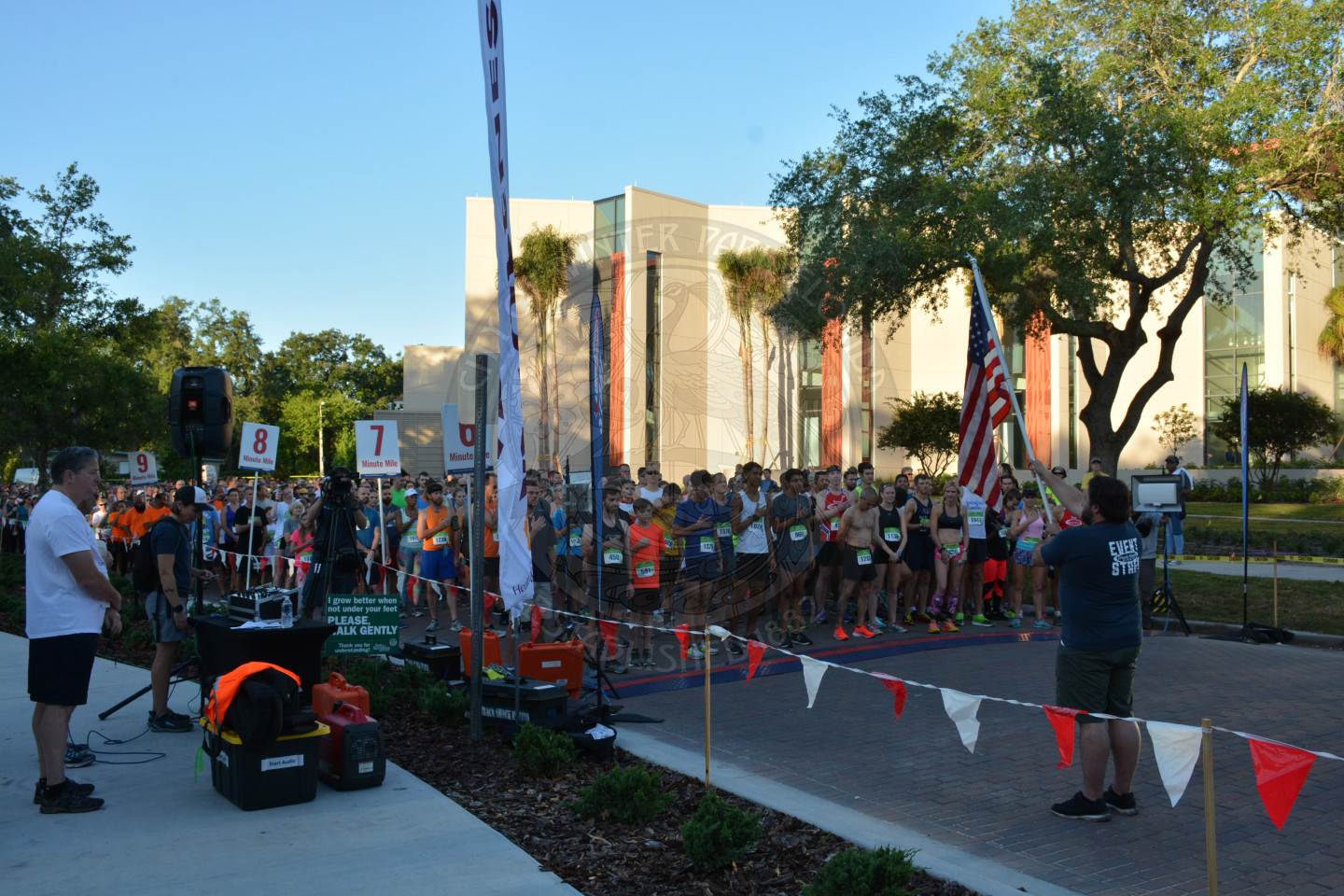 Runners and attendees reciting the Pledge of Allegiance with male holding the American Flag at the top of the race line.