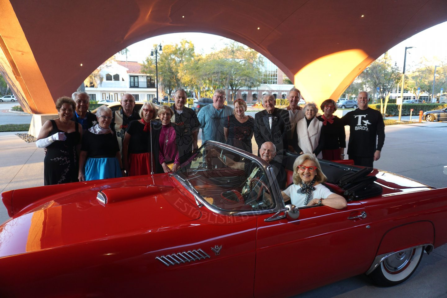 a women in the driver seat and a man in the passenger seat of a convertible with the hood down. There are people standing in the back of the convertible smiling. 