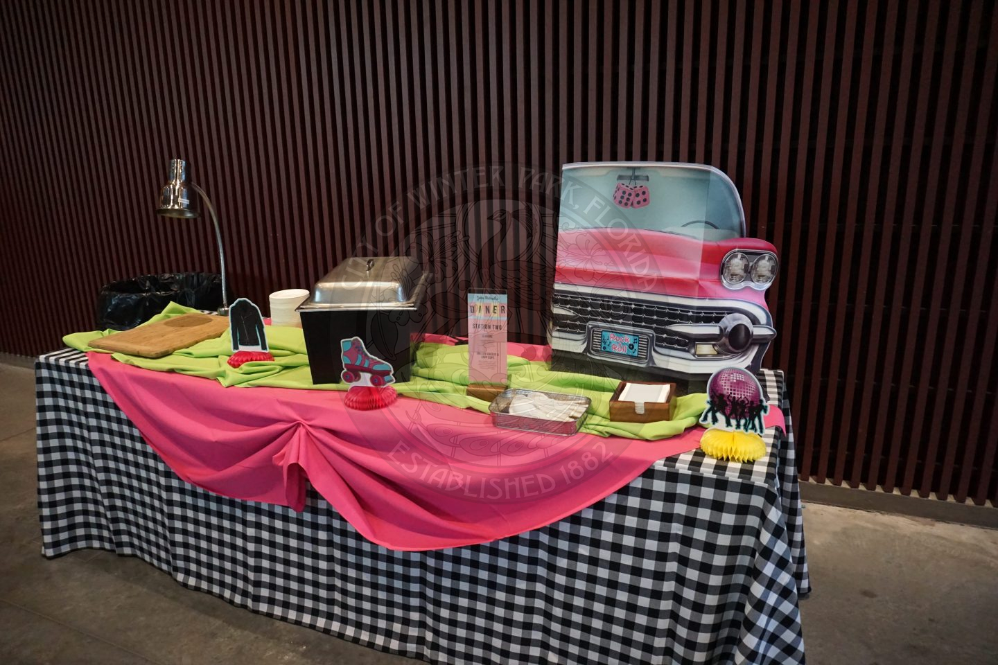 A dinner station with a black and white checkered table cloth, pink and green tableclothes on top of that. There is retro decor in between the hot pan with food.