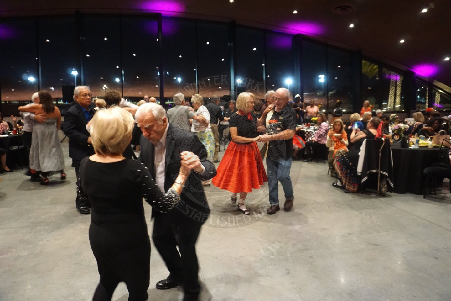 Senior couples holding each other and dancing in the middle of the dance floor. There are tables surounding the dance floor with some seniors sitting at the tables.