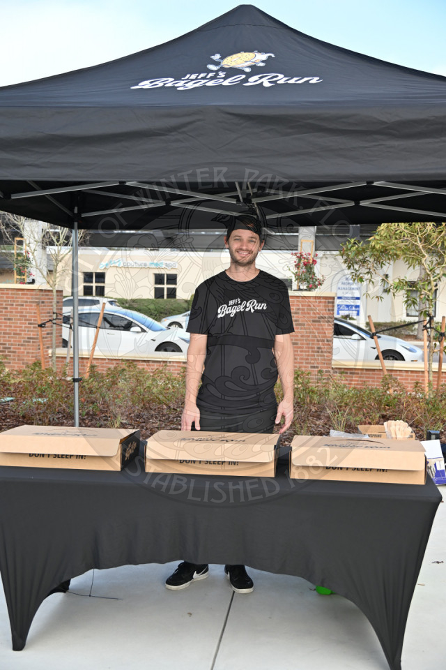 A guy standing behind a table with boxes of bagels.