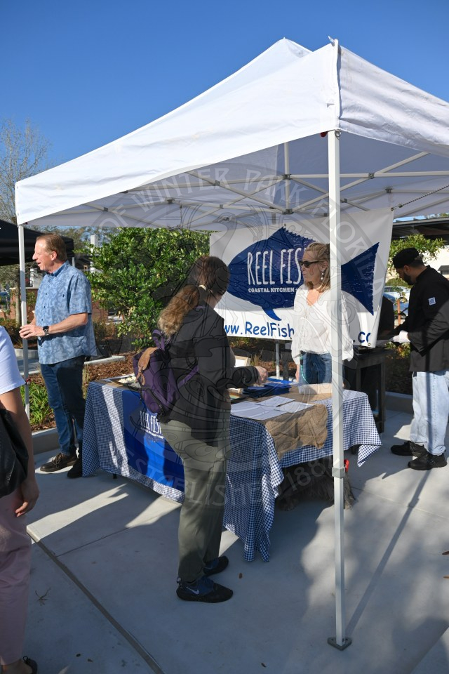 A white tent with the banner that says 'Reel Fish'. There is a table under the tent with infromation and people standing behind the table talking to people standing in front of the table.