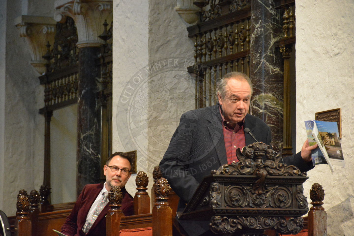 Gentleman standing at the podium sharing remarks while holding a copy of Winter Park Magazine. Reverend Shawn Garvey is sitting behind him attentively listening.