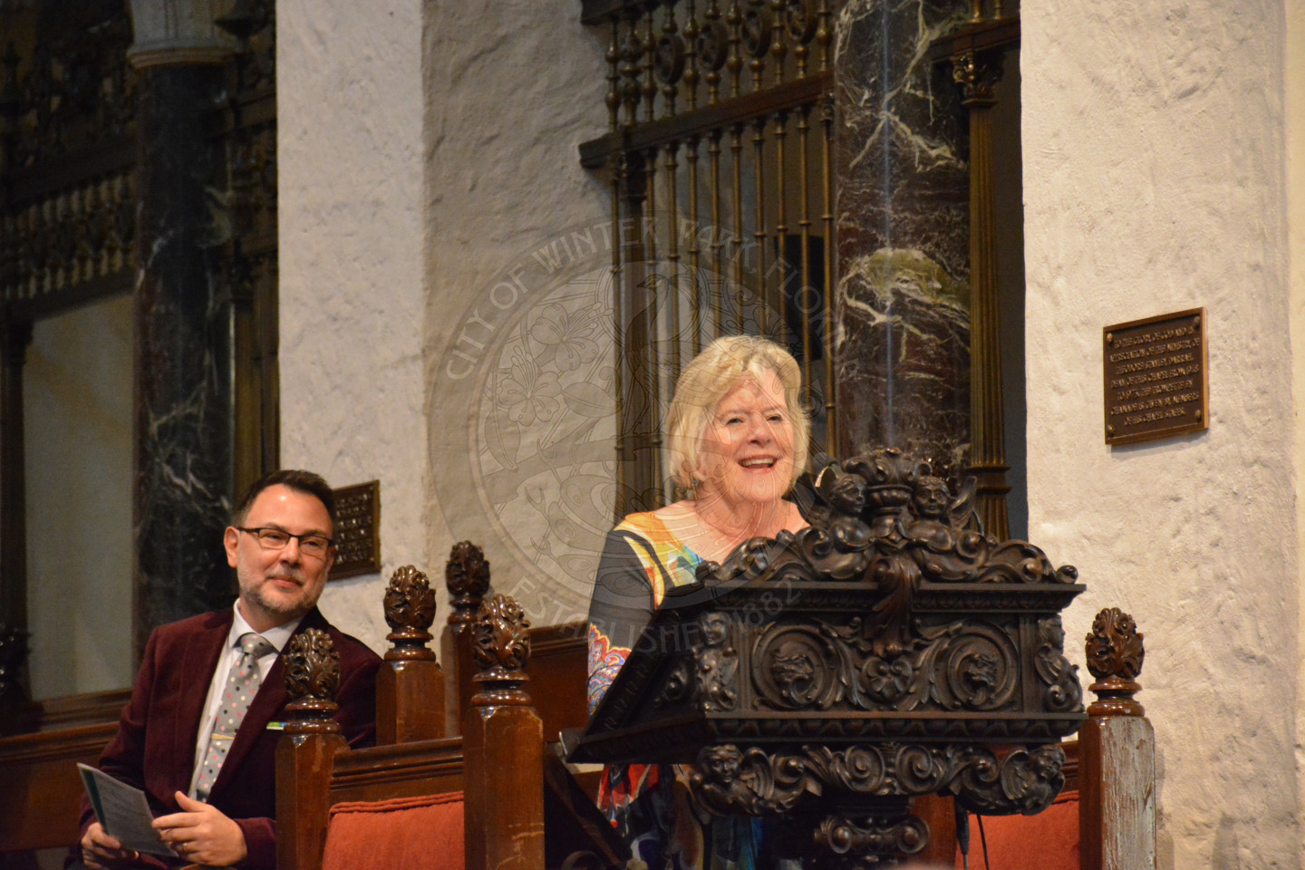 Female at the podium smiling while delivering remarks. Reverend Shawn Garvey is seated behind the speaker also smiling.