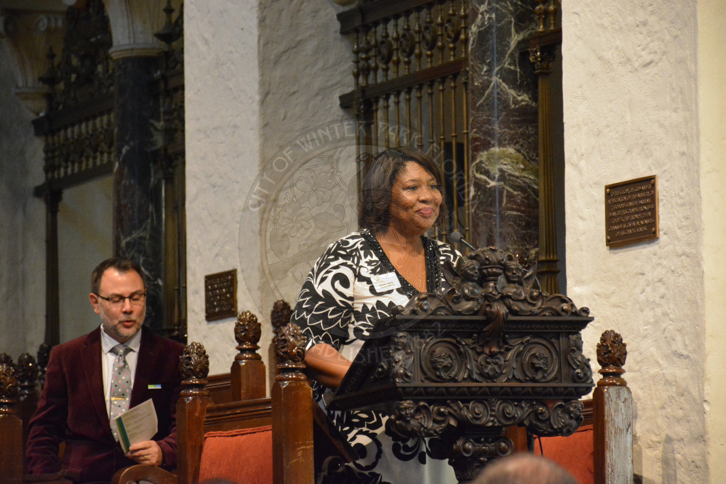 Female sharing remarks at the podium. Reverend Shawn Garvey seated behind the podium singing.