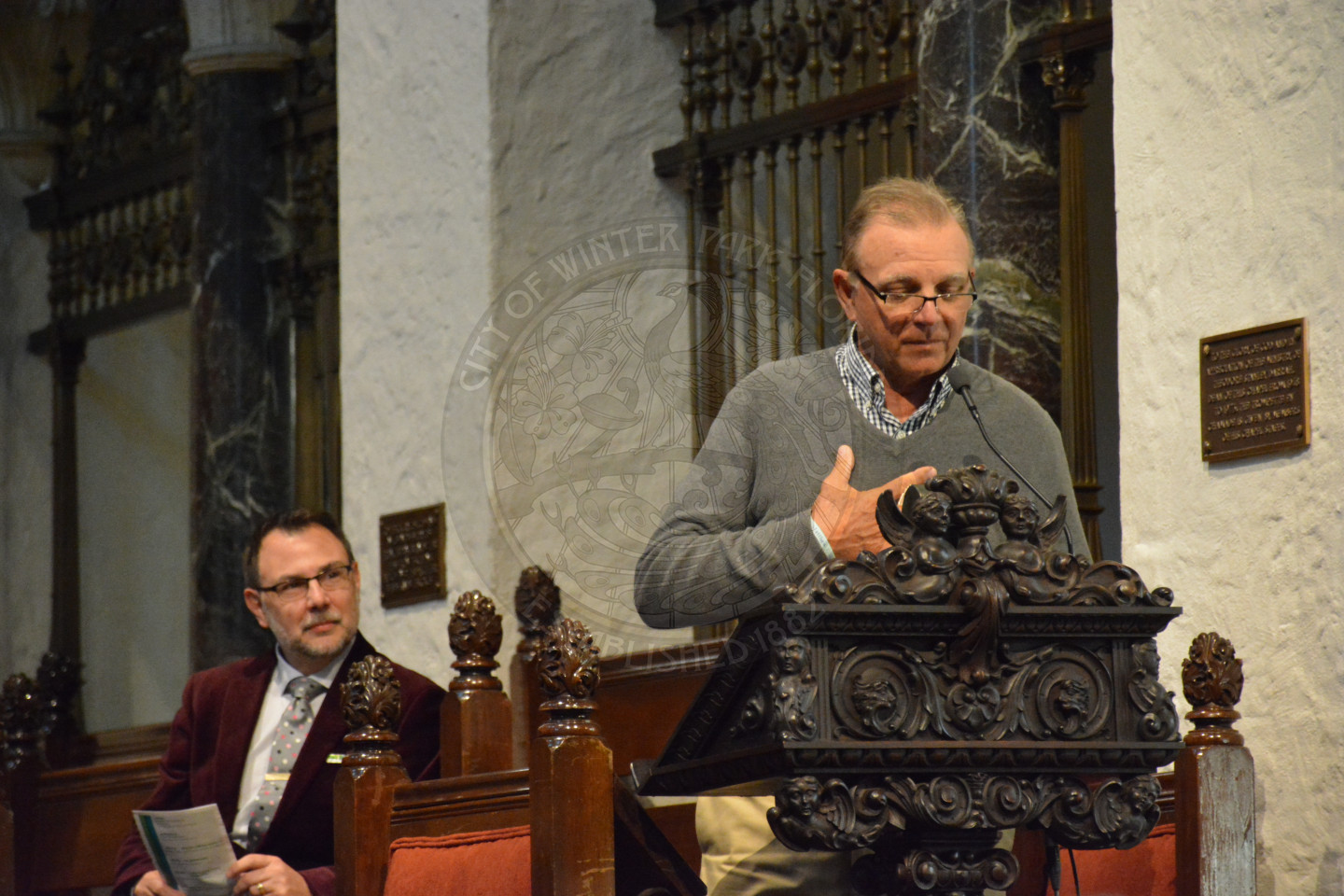 Gentleman at the podium sharing remarks. Reverend Shawn Garvey seated behind the podium listening.