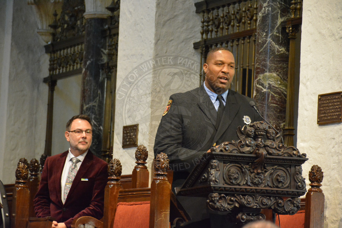 Winter Park Fire Rescue Ed Mitchell sharing remarks at the podium. Reverend Shawn Garvey is seated behind him.
