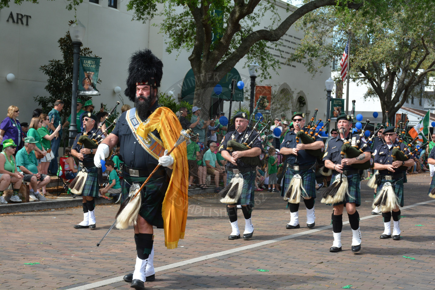 Bag pipers lead by man in tall furry hat, holding a long staff.