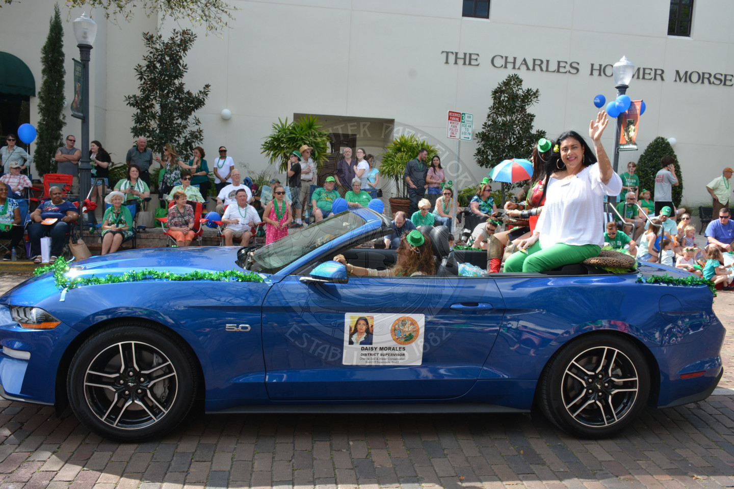 A female parade participant waving to parade attendees while sitting in a convertible vehicle.